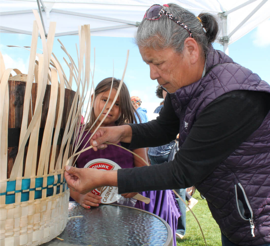home-Hero Image A woman and a child weave a basket together under a white canopy outdoors.