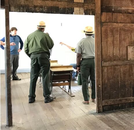People in conversation inside a rustic room, with two park rangers standing near wooden furniture.