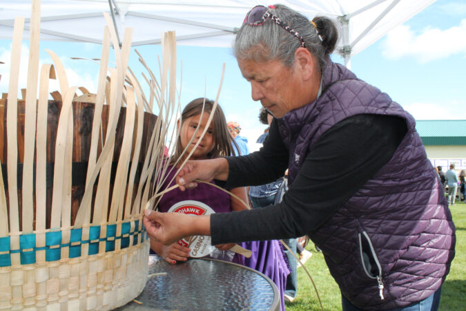 Two people weaving a large basket at an outdoor event under a tent.