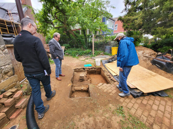 Three people examining a small excavation site in a backyard surrounded by greenery and brick walkways.