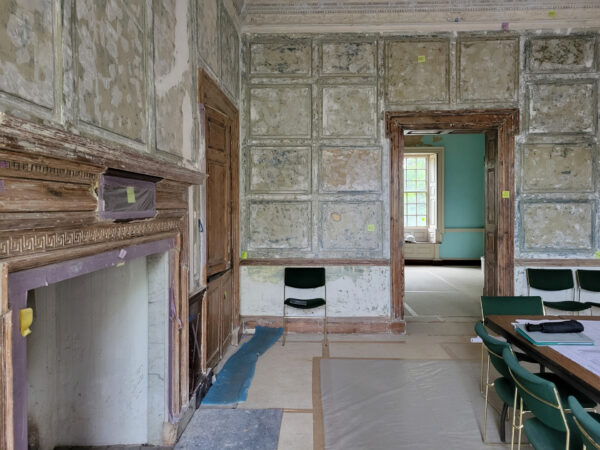 A room with peeling wall panels, a large fireplace, and a wooden table with chairs; an adjacent room is visible.