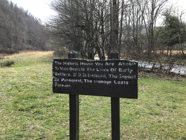 A wooden sign in a grassy field warns about defacing a historic house; trees and hills surround the area.