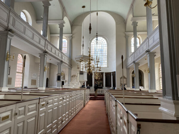 Interior of a historic church with white pews, tall columns, arched windows, and an ornate chandelier.