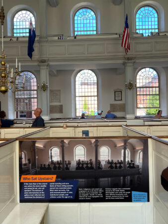 Interior of a historic church with large arched windows, informational display, and balcony seating.